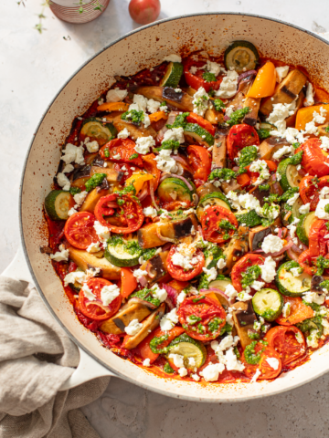 A close-up of a pan filled with colorful baked ratatouille—roasted tomatoes, zucchini, eggplant, and bell peppers—topped with crumbled cheese and green herb sauce on a light background.
