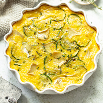 A baked zucchini frittata with melted chunks of brie, shown in a ruffle-edged baking dish.