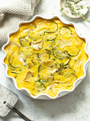 A baked zucchini frittata with melted chunks of brie, shown in a ruffle-edged baking dish.