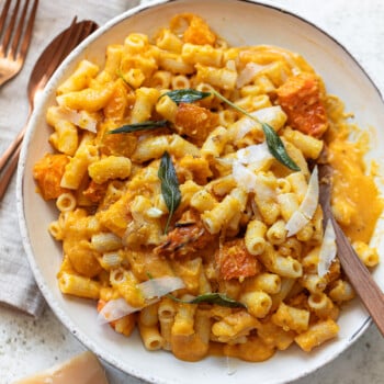 A bowl of butternut squash pasta with creamy orange sauce, chunks of roasted squash, shaved cheese, and fresh sage leaves, served with a wooden fork on a light surface next to a napkin, fork, and knife.