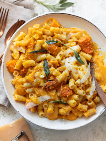 A bowl of butternut squash pasta with creamy orange sauce, chunks of roasted squash, shaved cheese, and fresh sage leaves, served with a wooden fork on a light surface next to a napkin, fork, and knife.