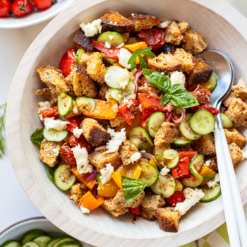 A wooden bowl filled with vibrant panzanella salad—toast cubes, cucumbers, cherry tomatoes, red onions, basil leaves, and crumbled cheese—with a spoon and fork resting inside.