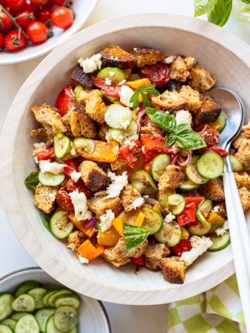 A wooden bowl filled with vibrant panzanella salad—toast cubes, cucumbers, cherry tomatoes, red onions, basil leaves, and crumbled cheese—with a spoon and fork resting inside.