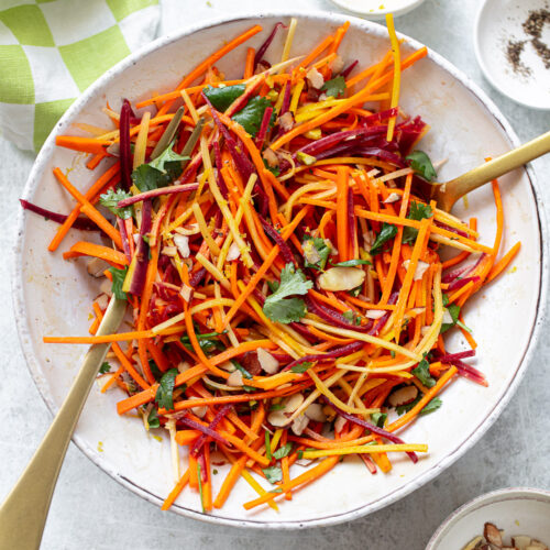 Matchstick-cut fresh carrot salad in a bowl, with serving utensils set alongside.