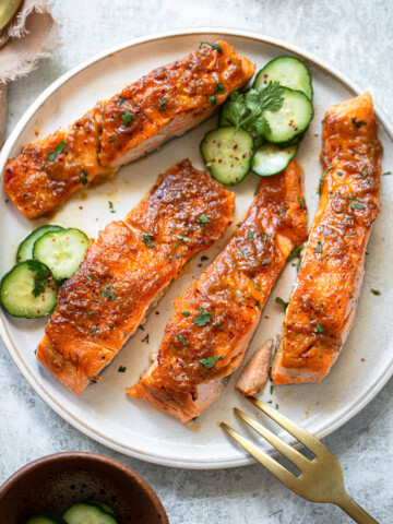 Maple glazed salmon on a serving plate, with sliced cucumbers and cilantro sprigs.