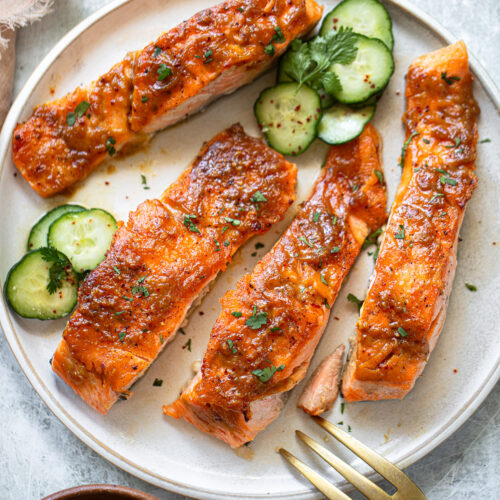 Maple glazed salmon on a serving plate, with sliced cucumbers and cilantro sprigs.