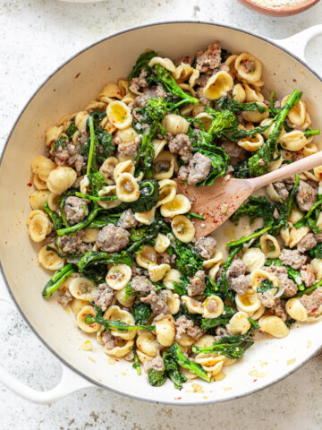 A white pot filled with orecchiette pasta, cooked sausage, and sautéed broccoli rabe—all mixed together for a delicious dinner. A wooden spoon rests inside the pot, ready to serve.