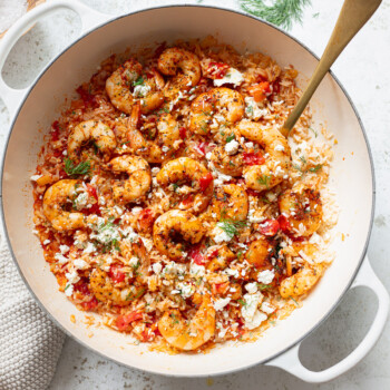 A white pot filled with savory shrimp and rice, tomatoes, herbs, and crumbled cheese, with a golden spoon resting inside. The dish is garnished with fresh dill and sits on a light-colored surface.