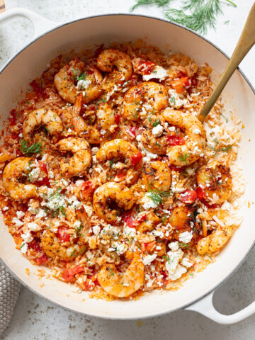 A white pot filled with savory shrimp and rice, tomatoes, herbs, and crumbled cheese, with a golden spoon resting inside. The dish is garnished with fresh dill and sits on a light-colored surface.