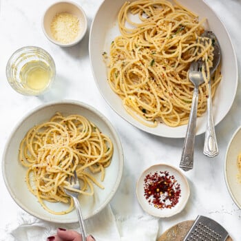 Spaghetti with garlic butter sauce on a platter, with a serving bowl on the side and a person's hand holding a fork and eating.