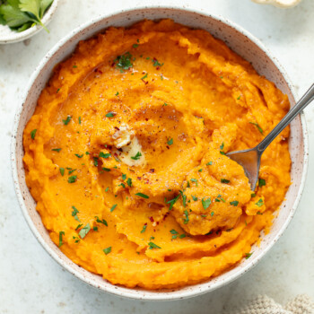 A bowl of creamy mashed butternut squash garnished with chopped herbs and pepper. A fork rests in the dish and a pat of melted butter tops it. The bowl sits on a light surface with parsley and a small dish nearby.