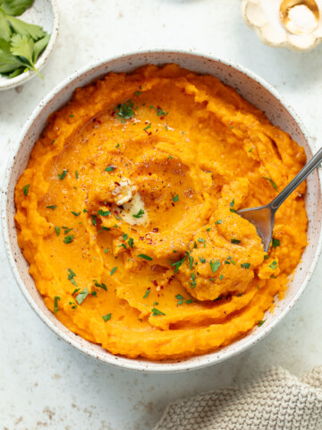 A bowl of creamy mashed butternut squash garnished with chopped herbs and pepper. A fork rests in the dish and a pat of melted butter tops it. The bowl sits on a light surface with parsley and a small dish nearby.