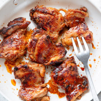 Six pieces of seared chicken thighs, browned to perfection with visible seasoning, sit in a white skillet with a fork resting beside them.
