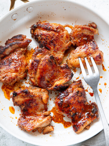 Six pieces of seared chicken thighs, browned to perfection with visible seasoning, sit in a white skillet with a fork resting beside them.
