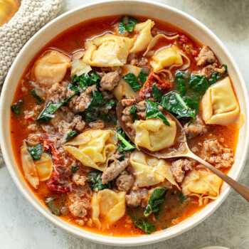 A spoon in a bowl of soup with chunky Italian sausage, tortellini pasta, and kale leaves.