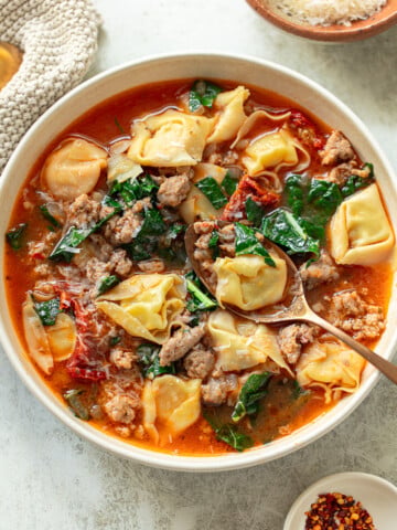 A spoon in a bowl of soup with chunky Italian sausage, tortellini pasta, and kale leaves.