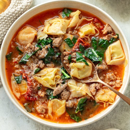 A spoon in a bowl of soup with chunky Italian sausage, tortellini pasta, and kale leaves.