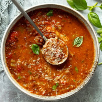 A bowl of hearty, tomato-based lentil soup with herbs, and grated cheese, topped with fresh basil leaves. A spoon rests in the bowl, scooping up a portion. o