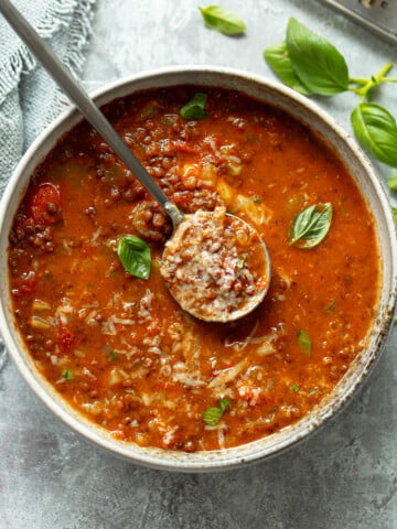 A bowl of hearty, tomato-based lentil soup with herbs, and grated cheese, topped with fresh basil leaves. A spoon rests in the bowl, scooping up a portion. o