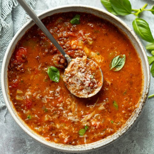 A serving bowl and spoon of lentil soup, with fresh basil and parsley sprinkled on top.
