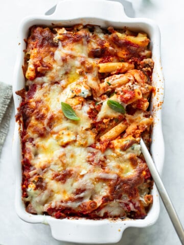 Ziti pasta and a serving spoon in a baking dish with melted cheese, marinara sauce and fresh basil leaves.