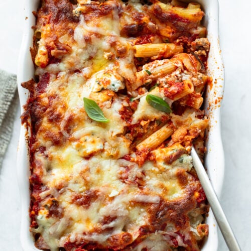 Ziti pasta and a serving spoon in a baking dish with melted cheese, marinara sauce and fresh basil leaves.