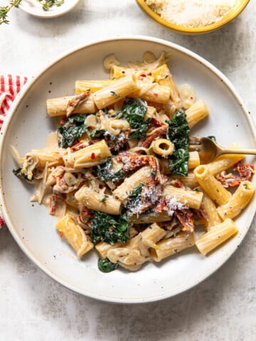 A plate of pasta with creamy sauce, sun-dried tomatoes, sautéed spinach, and grated cheese is served with a fork on a light surface. A striped napkin and small bowl of cheese are nearby.