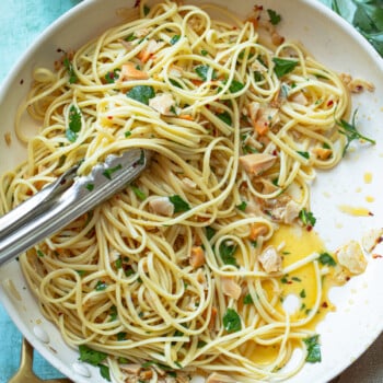 Linguine and clams in a white pan, mixed with garlic, parsley, and red pepper flakes. Olive oil is visible at the bottom, and kitchen tongs rest on top. A bowl of red pepper flakes is nearby on a light blue surface.