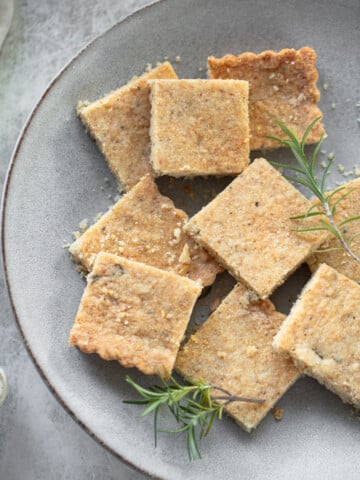 A gray plate with square-shaped shortbread biscuits, some with scalloped edges, sprinkled with crumbs. Fresh rosemary garnishes the arrangement, adding a touch of green. The background is a soft, textured gray surface.