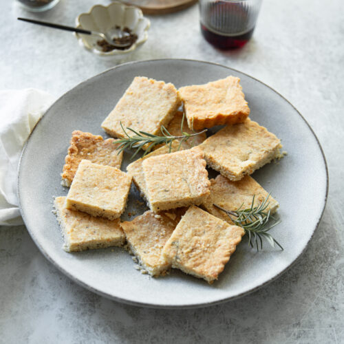 A plate of square cookies garnished with rosemary on a light gray surface. Nearby are a glass of red wine, a small jar of spices, and a wine bottle on a wooden coaster. A white cloth is also visible.
