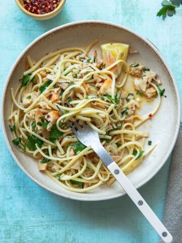 A plate of spaghetti with clams, garnished with parsley and chili flakes, sits on a light blue background. A lemon wedge rests on the plate. A white-handled fork twirls the pasta. A small bowl of chili flakes is in the upper corner.