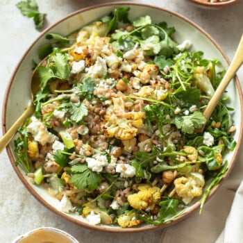 A bowl of roasted cauliflower salad with chickpeas, arugula, cilantro, and crumbled feta cheese placed for serving. Nearby are bowls of parsley and cilantro leaves, and a bowl of creamy dressing A napkin and wooden utensils rest beside the bowl on a light-colored surface.