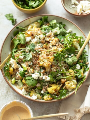 A bowl of roasted cauliflower salad with chickpeas, arugula, cilantro, and crumbled feta cheese placed for serving. Nearby are bowls of parsley and cilantro leaves, and a bowl of creamy dressing A napkin and wooden utensils rest beside the bowl on a light-colored surface.