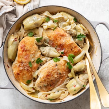 A top-down view of a skillet with golden-brown chicken breasts, artichoke hearts, and fettucchine pasta garnished with fresh parsley. A lemon slice and a napkin are beside the skillet, and two golden serving utensils rest inside.