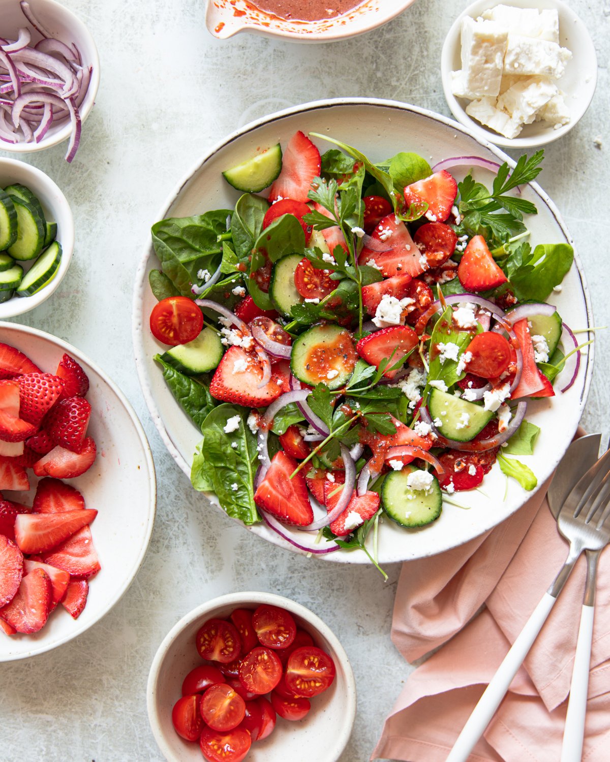 A fresh salad with spinach, strawberries, cherry tomatoes, cucumber slices, red onion, and feta cheese is on a plate. Surrounding are bowls of sliced strawberries, cherry tomatoes, feta, and dressing. A pink napkin with cutlery is nearby.