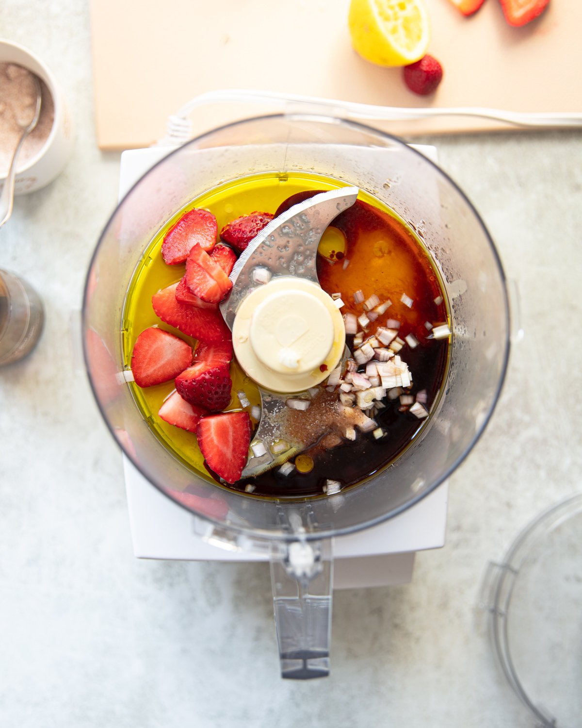 A top-down view of a food processor workbowl containing strawberries, chopped onions, olive oil, and balsamic vinegar. Ingredients are colorful against a light countertop, with a sliced lemon and strawberries nearby.