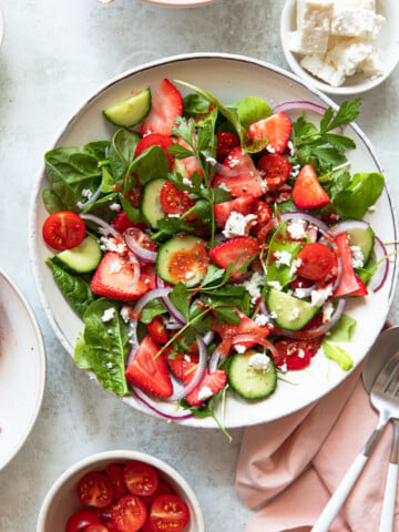 A vibrant salad with spinach, strawberries, cucumbers, cherry tomatoes, red onion, and parsley in a white bowl. Crumbled feta cheese is sprinkled on top. Surrounding dishes hold cherry tomatoes and feta. A pink napkin and fork are nearby.