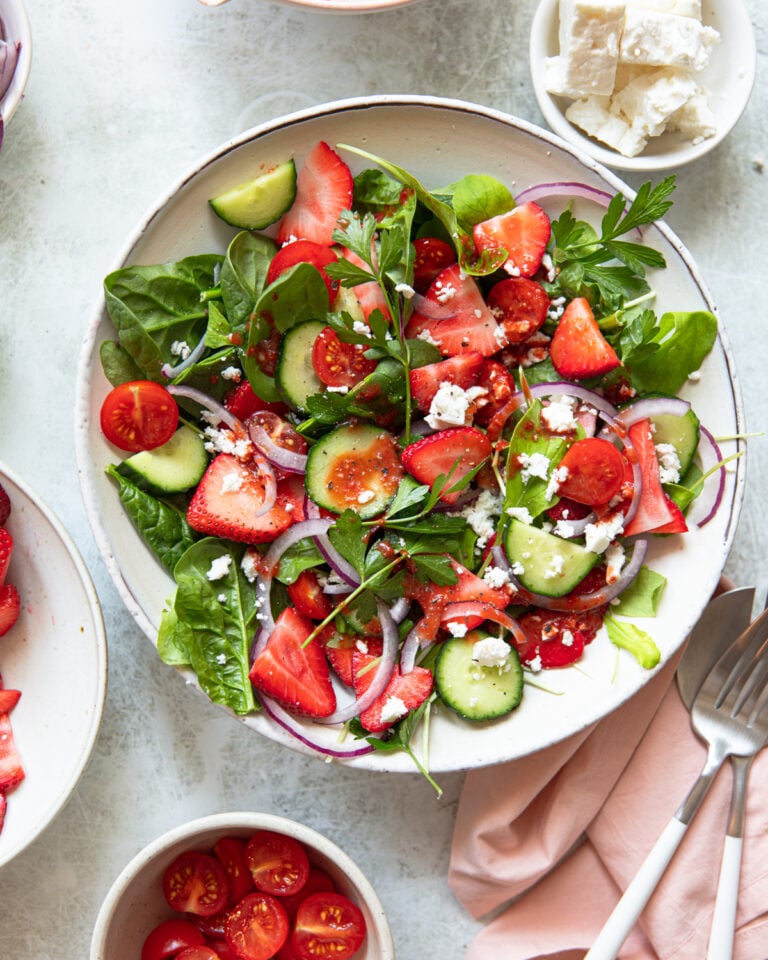 A vibrant salad with spinach, strawberries, cucumbers, cherry tomatoes, red onion, and parsley in a white bowl. Crumbled feta cheese is sprinkled on top. Surrounding dishes hold cherry tomatoes and feta. A pink napkin and fork are nearby.