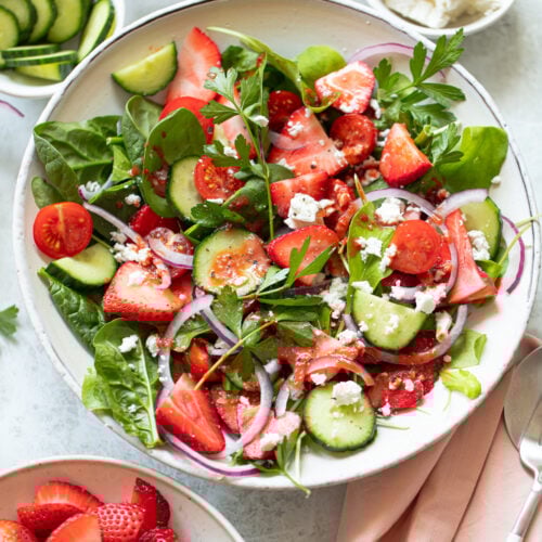 A fresh salad in a bowl featuring spinach, sliced strawberries, cucumbers, cherry tomatoes, red onion, and crumbled feta cheese. Garnished with parsley, the salad is colorful and vibrant. A small bowl of cucumbers and another of strawberries are nearby.