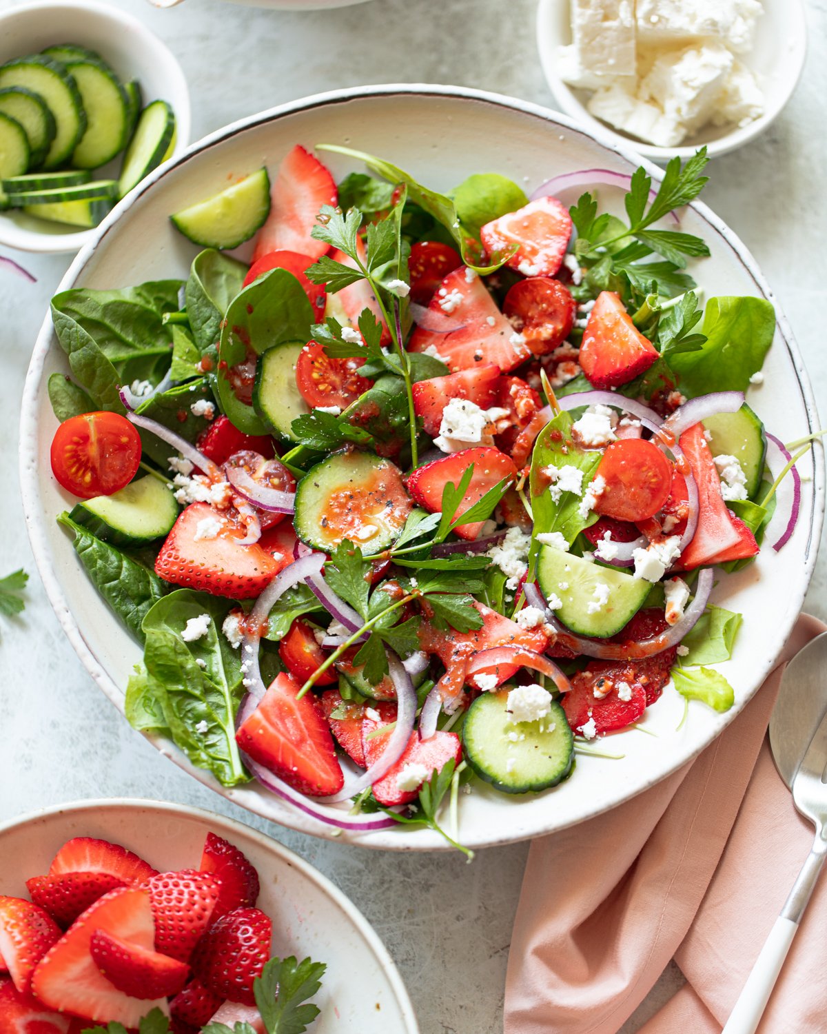 A fresh salad in a bowl featuring spinach, sliced strawberries, cucumbers, cherry tomatoes, red onion, and crumbled feta cheese. Garnished with parsley, the salad is colorful and vibrant. A small bowl of cucumbers and another of strawberries are nearby.