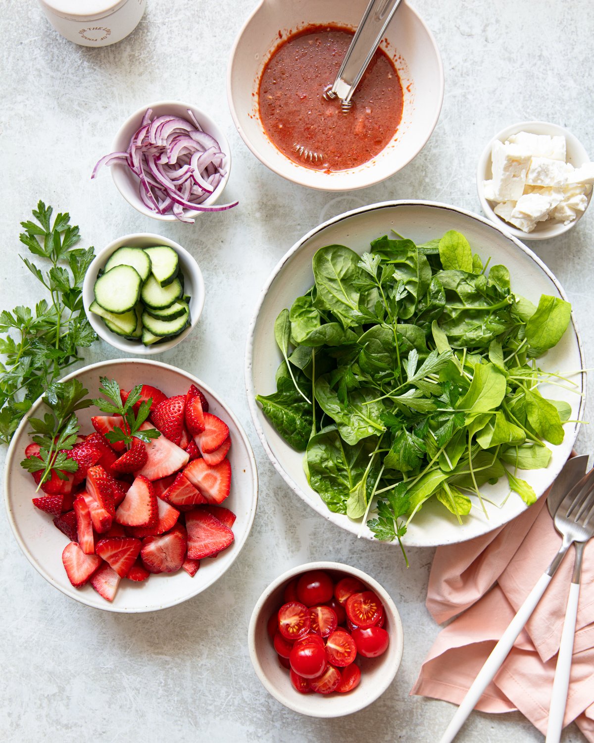 Assorted fresh salad ingredients including bowls of spinach, sliced strawberries, cherry tomatoes, cucumber slices, feta cheese, red onion, and a bowl of red dressing. A napkin and utensils are on the side.
