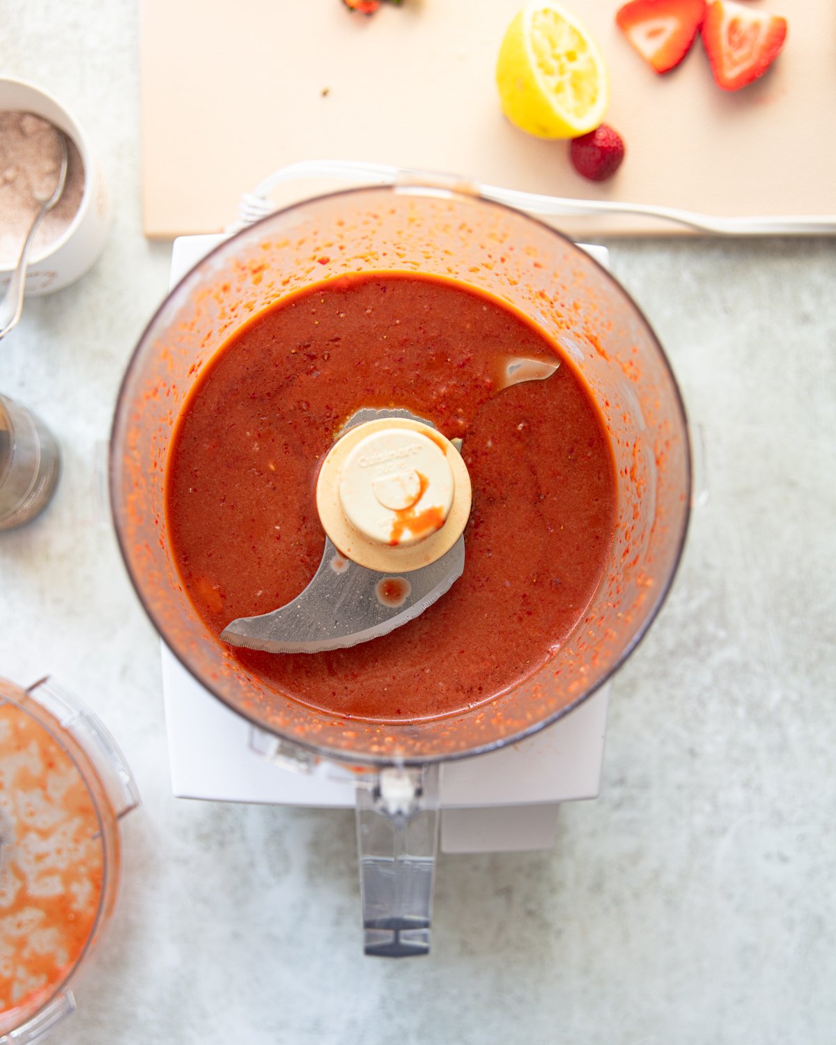 A food processor filled with blended strawberry vinaigrette sits on a light countertop. Nearby, a cutting board holds a halved lemon and fresh strawberries.