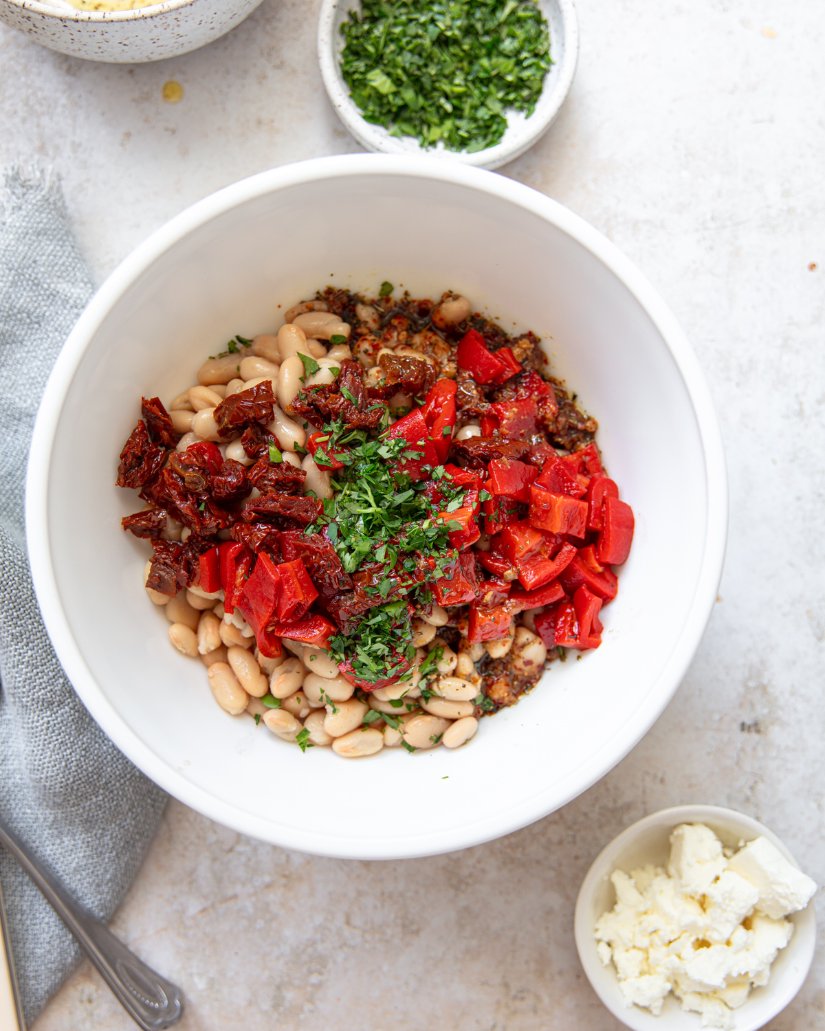 A white bowl filled with white beans, chopped red peppers, sun-dried tomatoes, herbs, and seasonings, surrounded by small bowls of feta cheese and chopped parsley on a light surface.