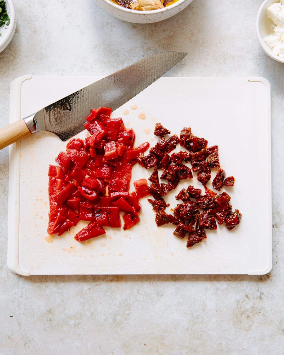 A cutting board with diced red bell peppers and chopped sun-dried tomatoes, next to a kitchen knife. Small bowls with ingredients are visible around the board on a light countertop.