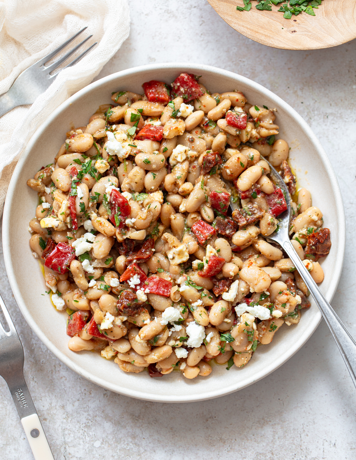 A bowl of white bean salad with chopped red peppers, herbs, and crumbled cheese, served with a fork on a light surface. A napkin and another fork are nearby.