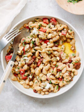 A white bowl filled with a white bean salad, mixed with chopped red peppers, herbs, and crumbled cheese. A fork and spoon rest on the bowl’s edge, and a drizzle of olive oil is visible.
