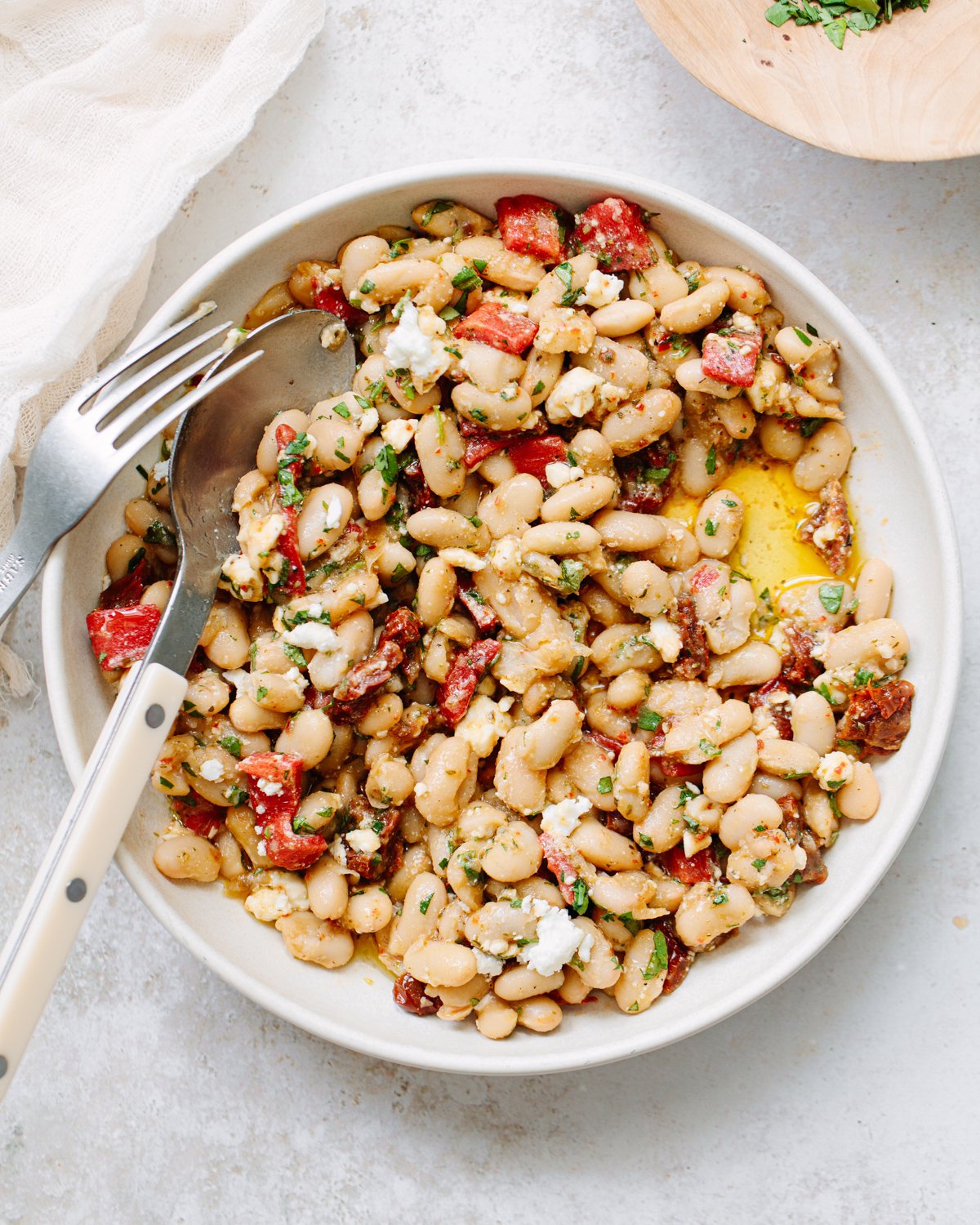 A white bowl filled with a white bean salad, mixed with chopped red peppers, herbs, and crumbled cheese. A fork and spoon rest on the bowl’s edge, and a drizzle of olive oil is visible.