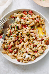 A bowl of white bean salad with chopped herbs, crumbled cheese, sun-dried tomatoes, and diced red peppers, drizzled with olive oil. A fork and spoon rest on the edge of the bowl.