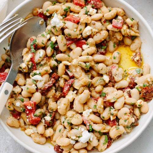 A bowl of white bean salad with chopped herbs, crumbled cheese, sun-dried tomatoes, and diced red peppers, drizzled with olive oil. A fork and spoon rest on the edge of the bowl.