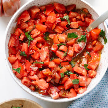A bowl of freshly chopped tomatoes mixed with basil and olive oil, with a spoon inside the bowl. A whole tomato, basil leaves, and a blue cloth are nearby on a white surface.